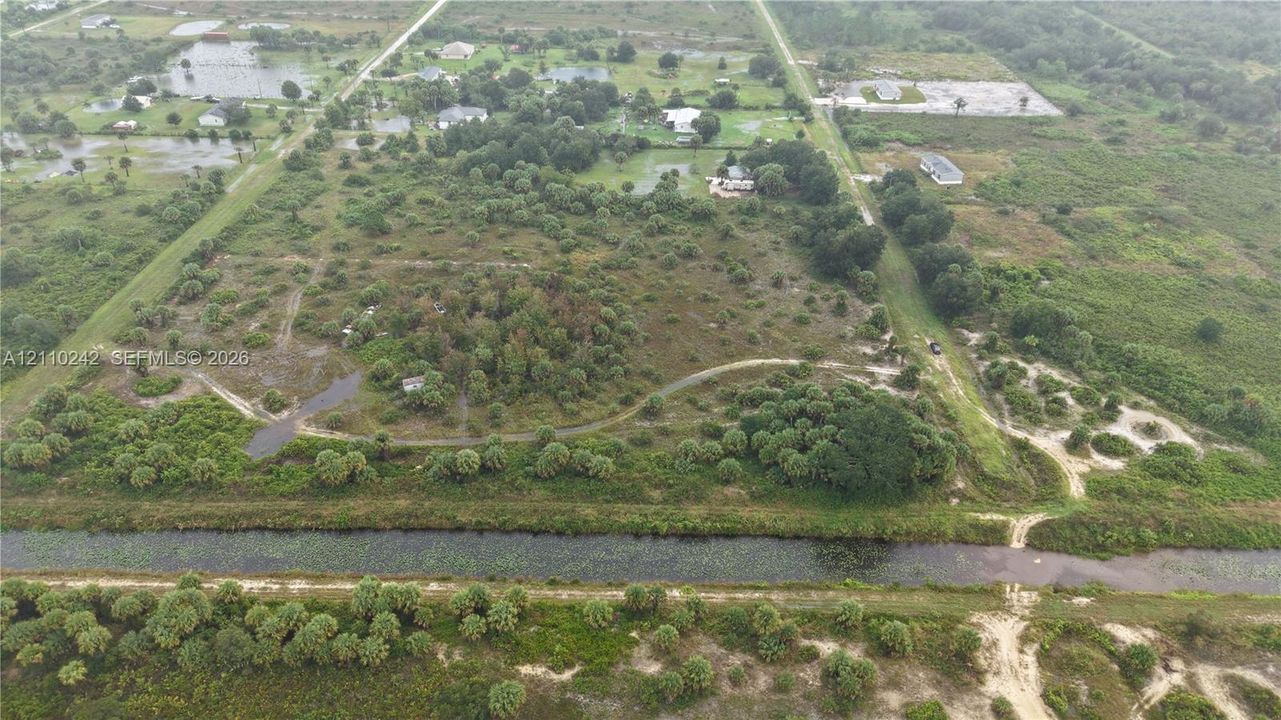 Expansive land parcel featuring native vegetation, clusters of mature trees, unpaved access paths, and a bordering canal