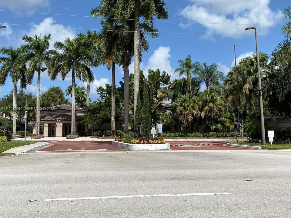 Paved entry gatehouse featuring red brick-patterned driveway, mature palm trees, landscaped center island, and surrounding tropical foliage
