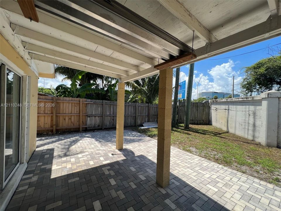 Covered patio featuring a paver surface, structural block columns, and an exposed beam ceiling, leading to a fenced backyard