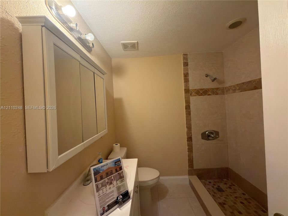 Bathroom featuring a built-in medicine cabinet with integrated vanity lighting, tiled walk-in shower with decorative stone-finish accents, and tiled flooring