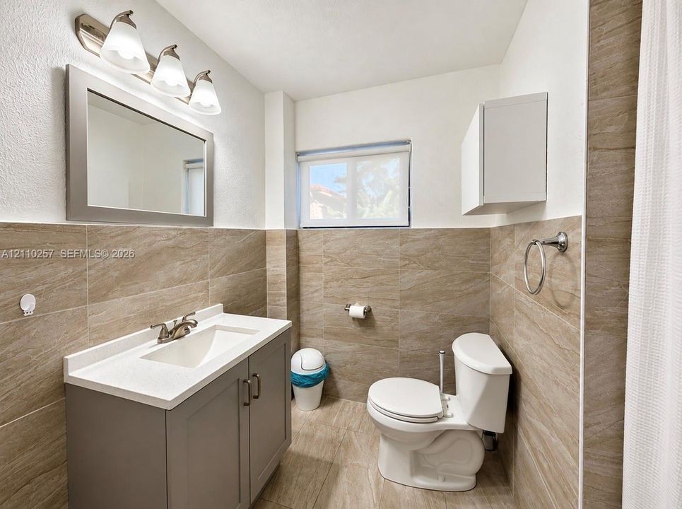 Bathroom featuring stone-finish wall and floor tiling, vanity with a light-toned countertop, framed wall mirror, three-light vanity fixture, and white wall-mounted cabinetry