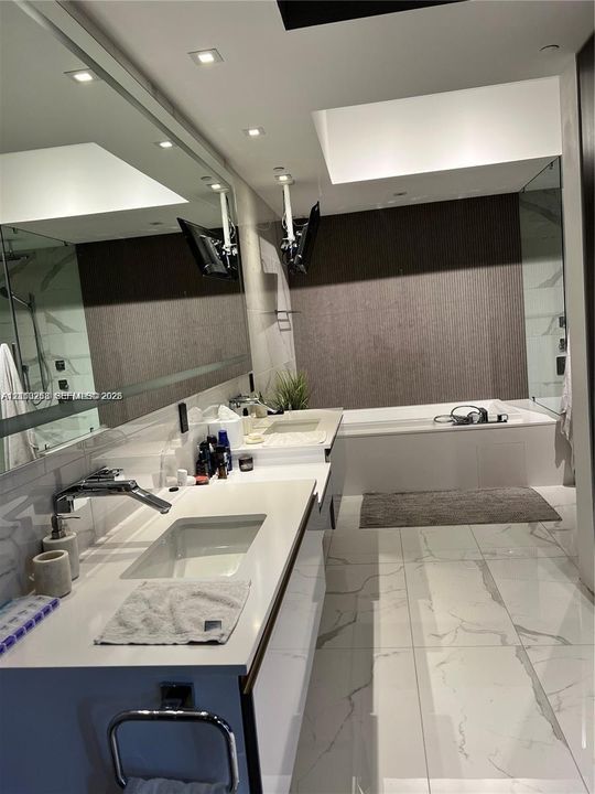 Modern bathroom featuring a double vanity with integrated rectangular sinks, white countertop, full-width mirror, recessed ceiling lighting, and a deep soaking tub with textured wall surround