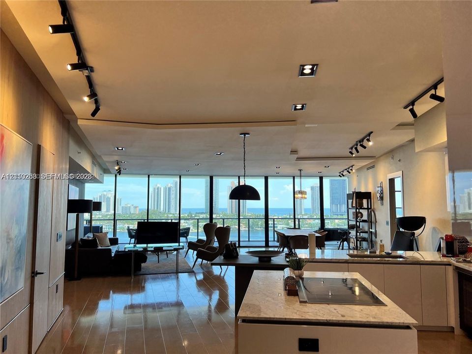 Open-concept living space featuring floor-to-ceiling windows, wood-finish flooring, a kitchen island with an integrated cooktop, stone-surfaced countertops, and black track lighting