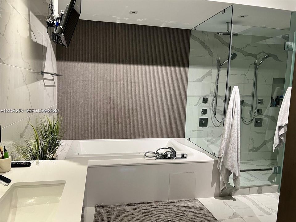 Soaking tub with a white composite surround, walk-in glass-enclosed shower featuring dual showerheads, textured accent wall, and stone-finish wall and floor tile