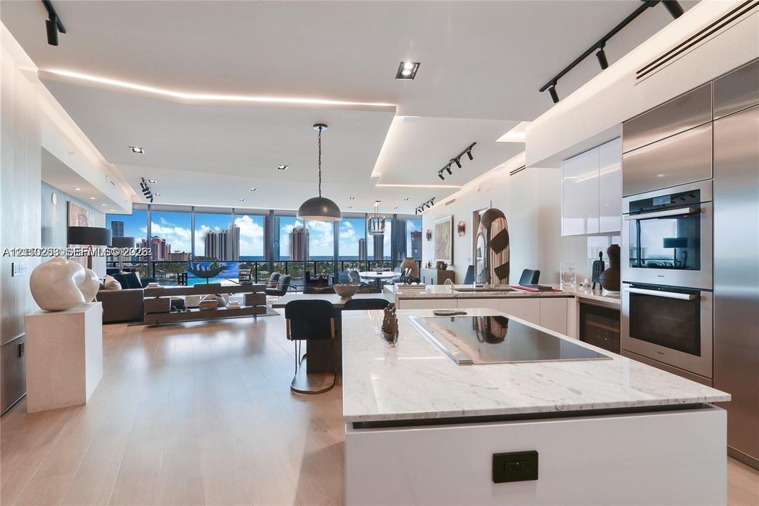 Modern kitchen featuring a central island with integrated cooktop and stone-surface countertop, floor-to-ceiling windows with urban skyline backdrop, wood-finish flooring, built-in stainless steel double wall ovens, and custom recessed lighting