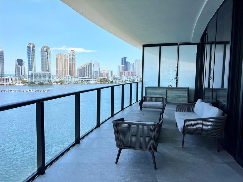 Expansive covered balcony featuring large-format stone flooring, black metal railing, and floor-to-ceiling glass wall panels