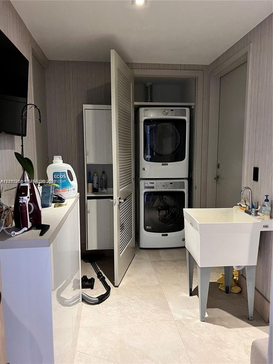 Laundry area featuring stacked white washer and dryer, built-in white cabinetry, louvered closet door, freestanding utility sink with chrome faucet, and tile flooring