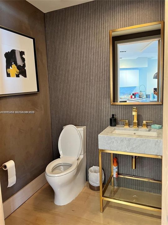 Textured accent wall featuring a wall-mounted console vanity with stone countertop, integrated sink, brass-finish faucet, and coordinating brass-framed mirror
