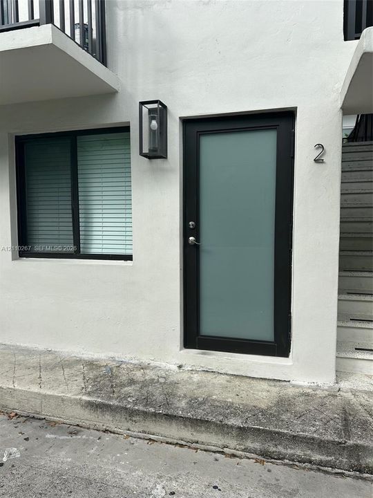 Exterior entrance featuring a white stucco facade, a black-framed door with a frosted glass panel, a sleek rectangular wall-mounted light fixture, and a black-framed window
