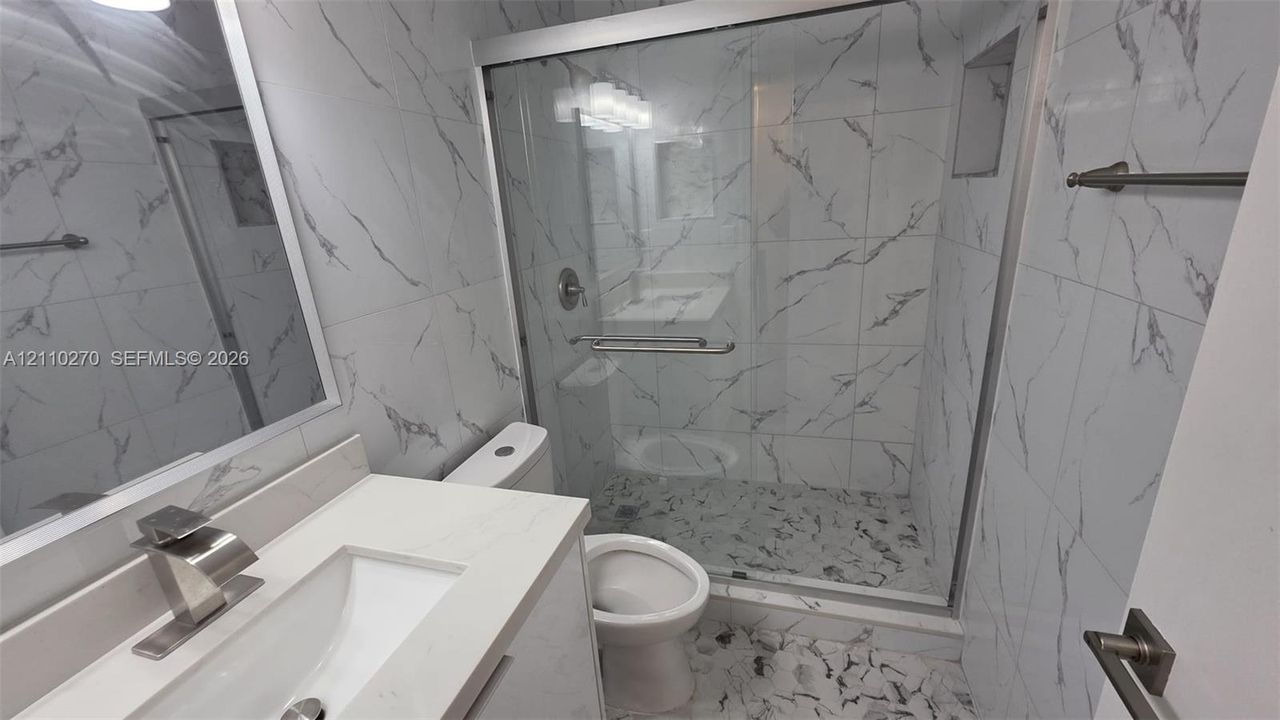 Bathroom featuring large-format wall tiling with a veined pattern, glass-enclosed shower with pebble-stone flooring, white vanity with a composite countertop, rectangular undermount sink, and brushed-finish metal fixtures