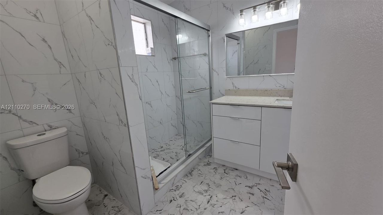 Bathroom featuring large-format wall tiles, glass-enclosed shower, stone-finish vanity countertop, and rectangular wall mirror with multi-bulb vanity lighting