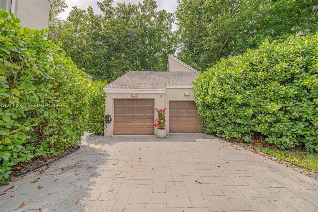 Detached two-car garage with paneled doors, expansive stone-paver driveway, and mature perimeter landscaping
