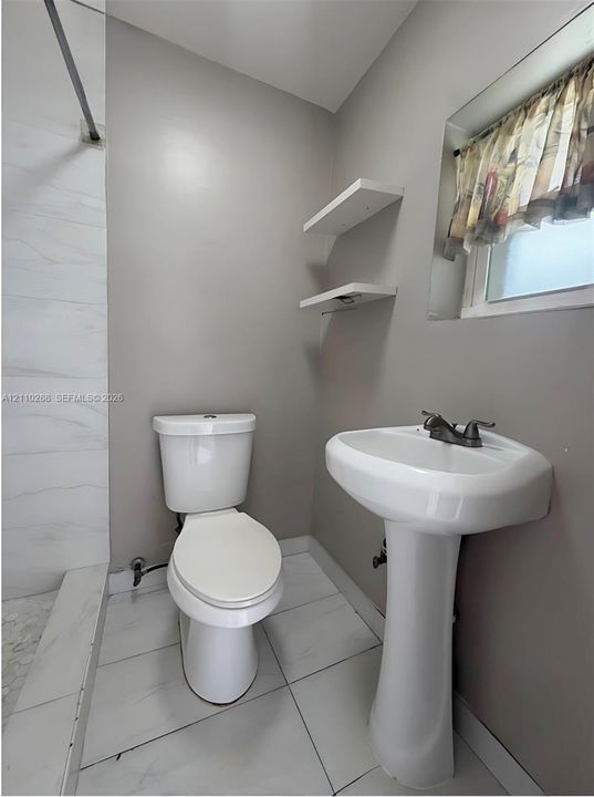 Bathroom featuring a white pedestal sink, dual-handle faucet, white elongated toilet, tiled flooring, and built-in wall shelving