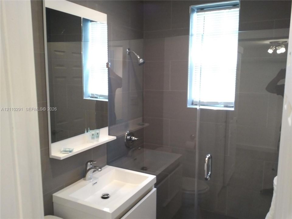 Modern floating vanity with a rectangular sink, wall-mounted mirror with integrated shelf, glass-enclosed shower, polished chrome fixtures, and grey ceramic wall tiling