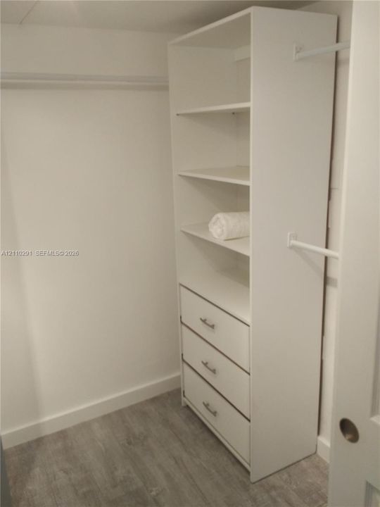Custom closet organizer featuring white shelving, three built-in drawers with metallic hardware, and white hanging rods, set against wood-finish flooring