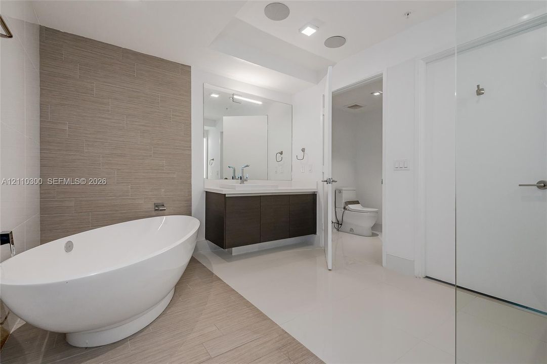 Master Bathroom Freestanding oval soaking tub, wall-mounted faucet, floating wood-finish vanity with a rectangular sink, large format tile flooring, and textured tile accent wall