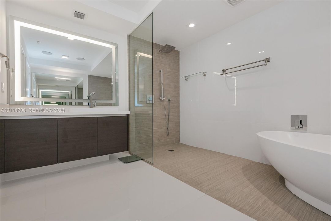 Master Bathroom Floating vanity with dark wood-finish cabinetry, integrated LED-backlit mirror, walk-in glass shower, wall-mounted shower hardware, and freestanding soaking tub