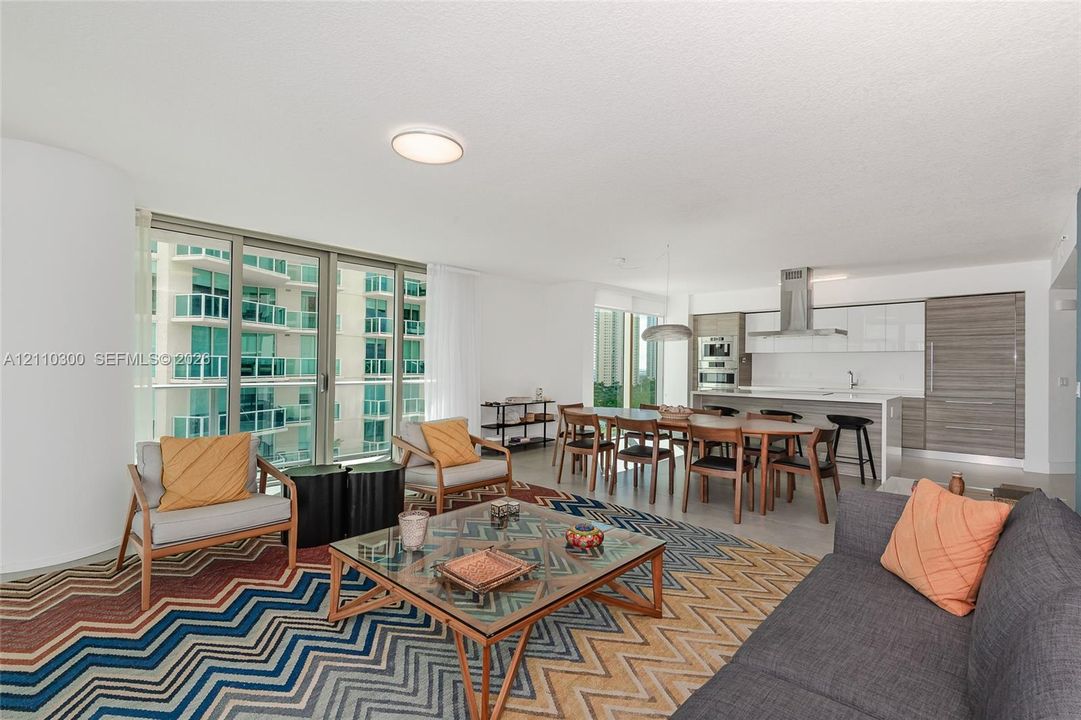 Open-concept living space featuring floor-to-ceiling glass sliding doors, modern cabinetry with a dual-tone finish, built-in stainless steel appliances, a kitchen island with stone-finish countertops, and large-format tile flooring
