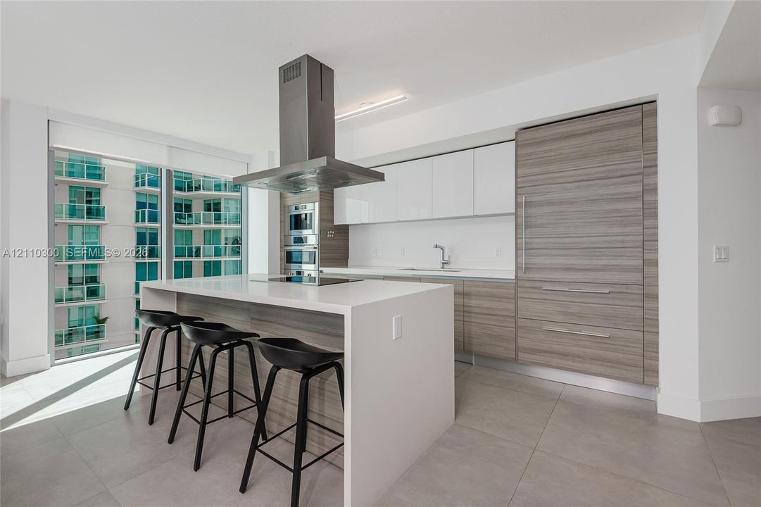 Modern kitchen featuring an island with a waterfall-edge countertop, panel-ready refrigerator, stainless steel ceiling-mounted range hood, built-in double wall ovens, and two-tone flat-panel cabinetry
