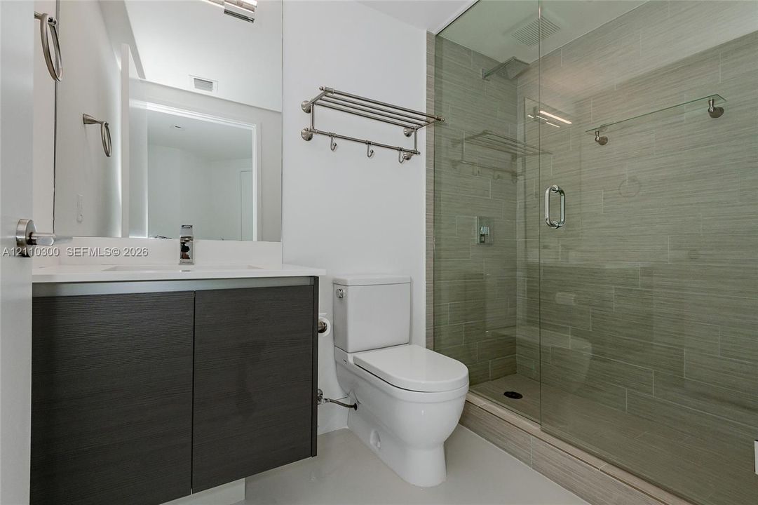 Bathroom 3. featuring a vanity with wood-finish cabinetry and a white countertop, walk-in shower with a glass enclosure and textured tile surround, wall-mounted metal shelving, and a dual-flush toilet