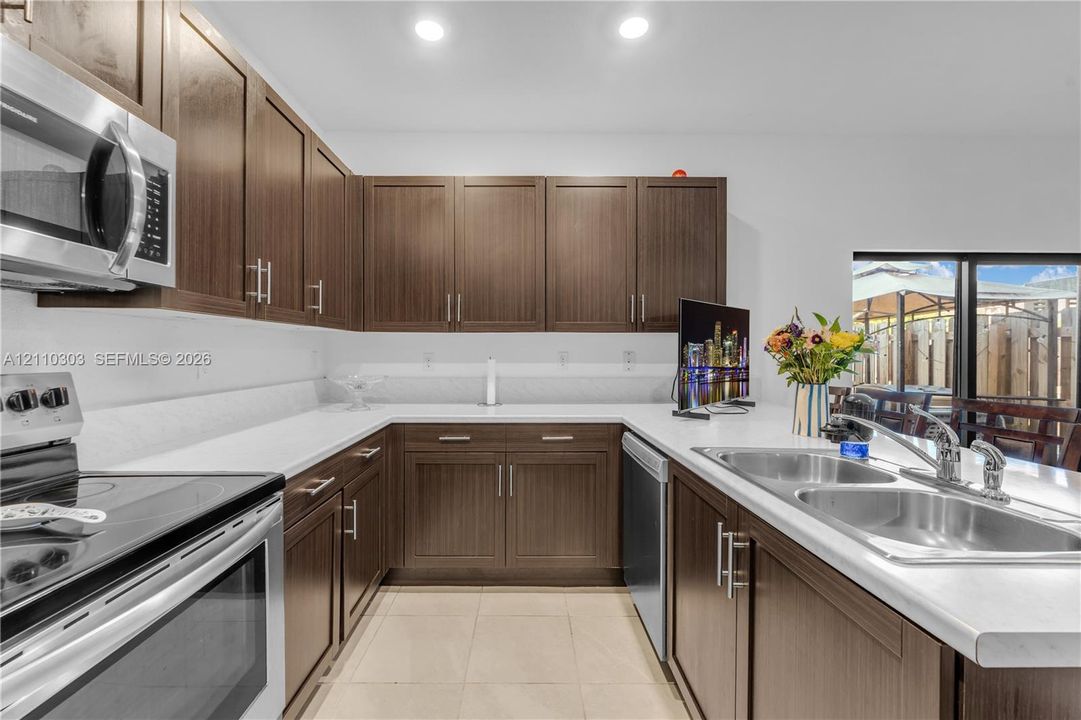 Kitchen featuring dark wood-finish cabinetry, solid surface countertops, stainless steel dual-basin sink, integrated stainless steel appliances, and tile flooring