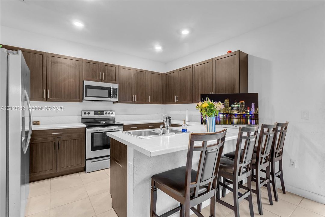 Kitchen featuring dark wood-finish cabinetry, white countertops, a central island with a double-basin sink, and stainless steel appliances including a built-in microwave and range