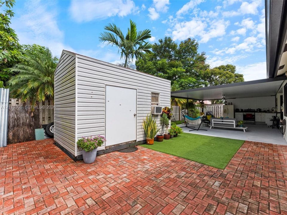 Rear exterior space featuring a herringbone-patterned brick patio, synthetic turf area, covered lanai, and detached storage structure