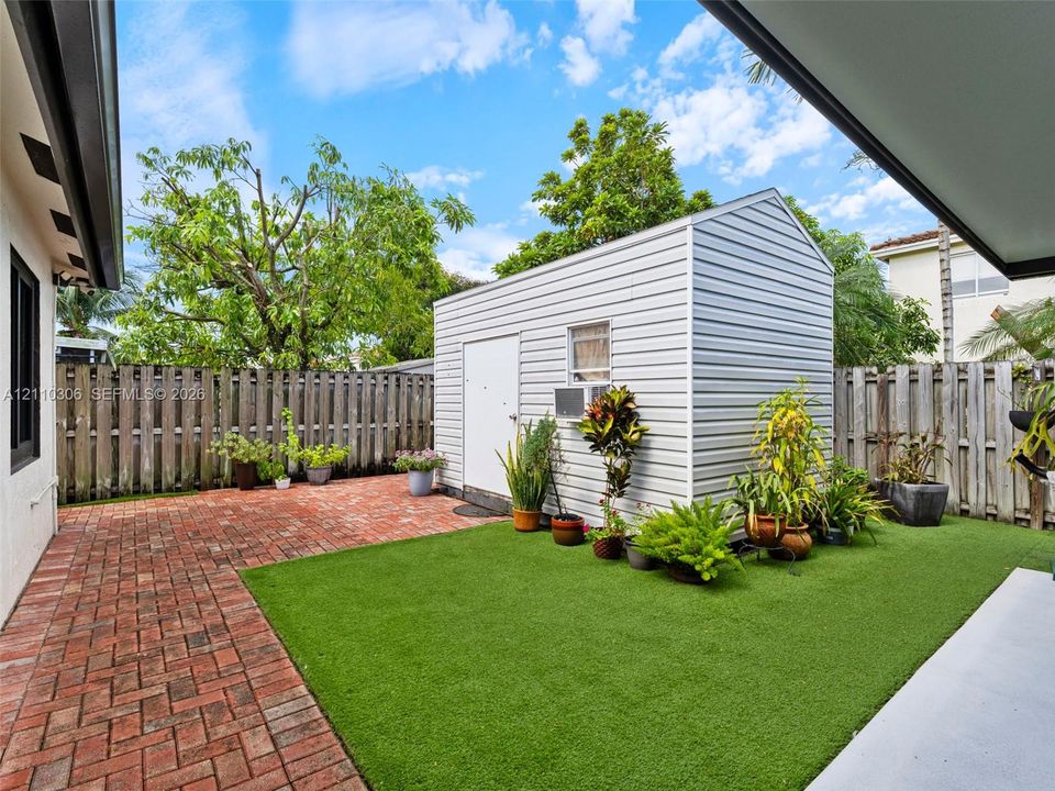 Backyard featuring red brick hardscaping, artificial turf lawn, privacy fencing, and a detached metal storage structure