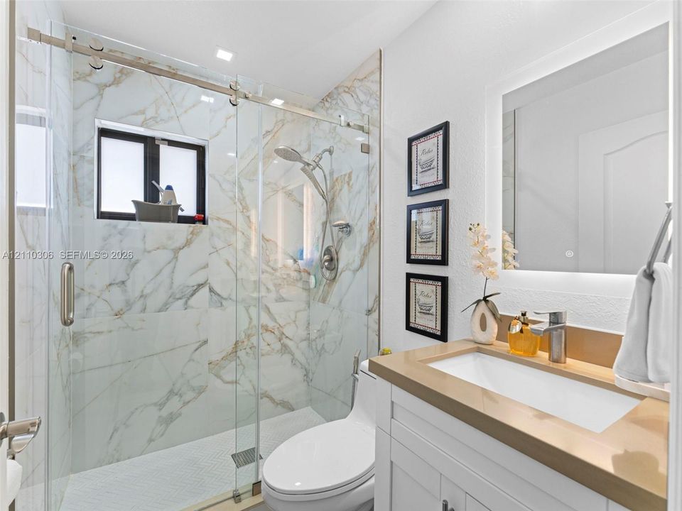 Walk-in shower with floor-to-ceiling veined tile, sliding glass enclosure, rainfall shower head, white vanity with integrated basin, and backlit wall mirror