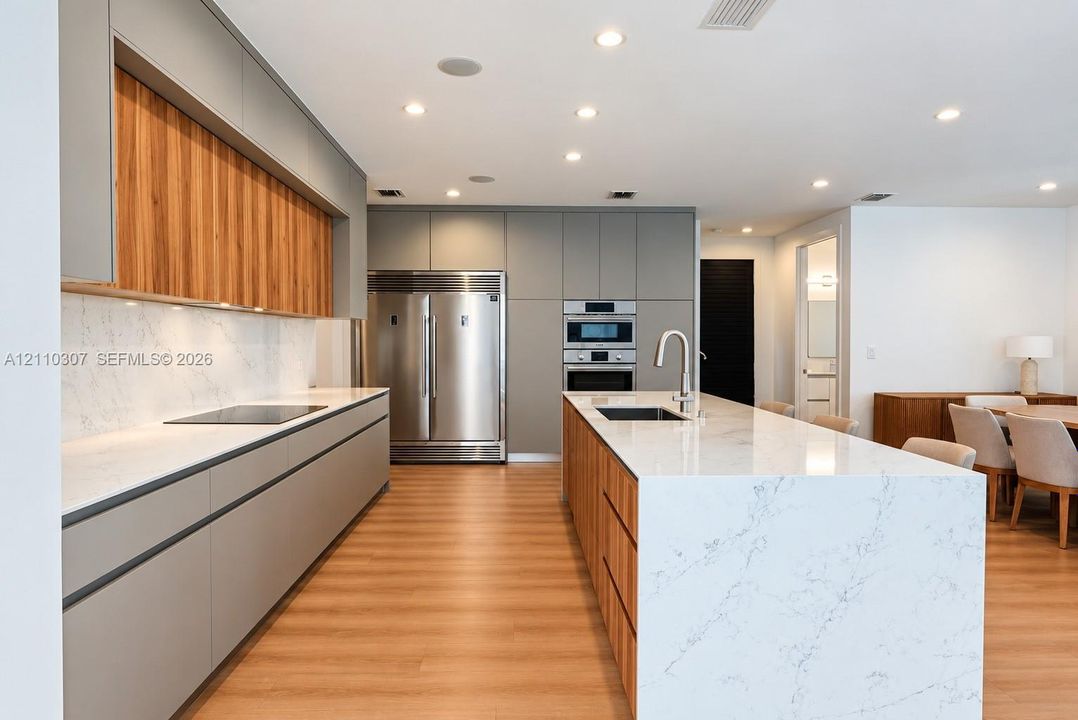 Modern kitchen featuring two-tone flat-panel cabinetry, a central island with a waterfall countertop, integrated stainless steel appliances, and wood-finish flooring