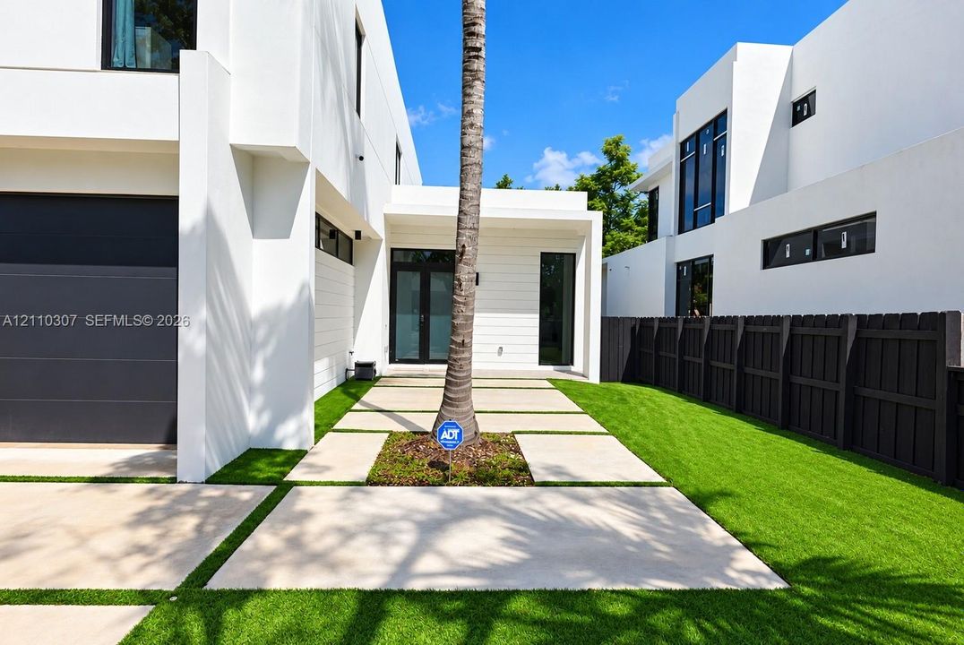 Modern architectural exterior featuring smooth white stucco finish, geometric paver pathway, synthetic turf lawn, dark-toned panel garage door, and tall black-framed windows