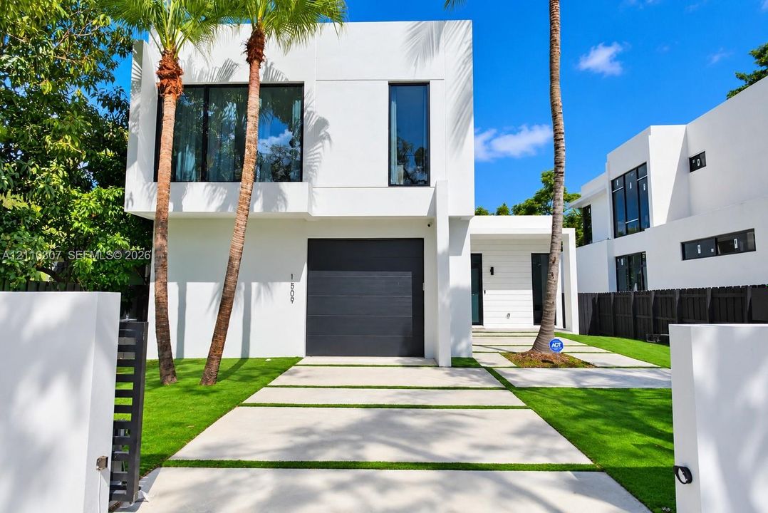 Modern two-story architectural exterior featuring smooth white stucco finish, large dark-framed windows, a sleek charcoal-colored garage door, and a geometric concrete paver driveway
