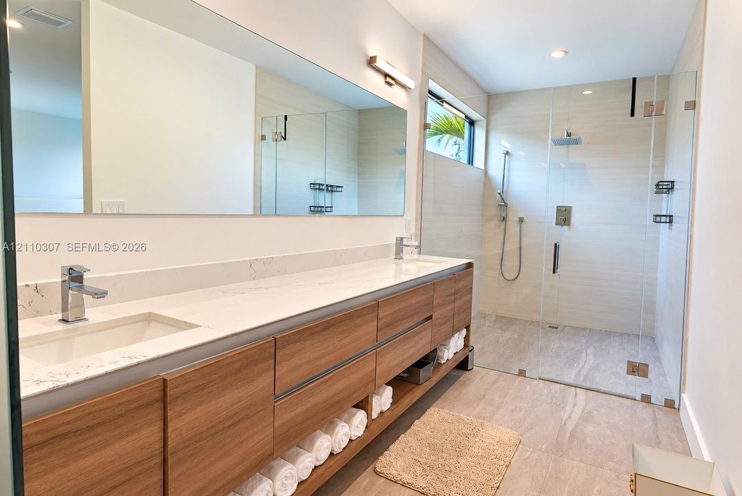 Dual-sink vanity with wood-finish cabinetry, light-toned stone-finish countertop, full-width wall mirror, and walk-in shower with glass enclosure and rainfall showerhead