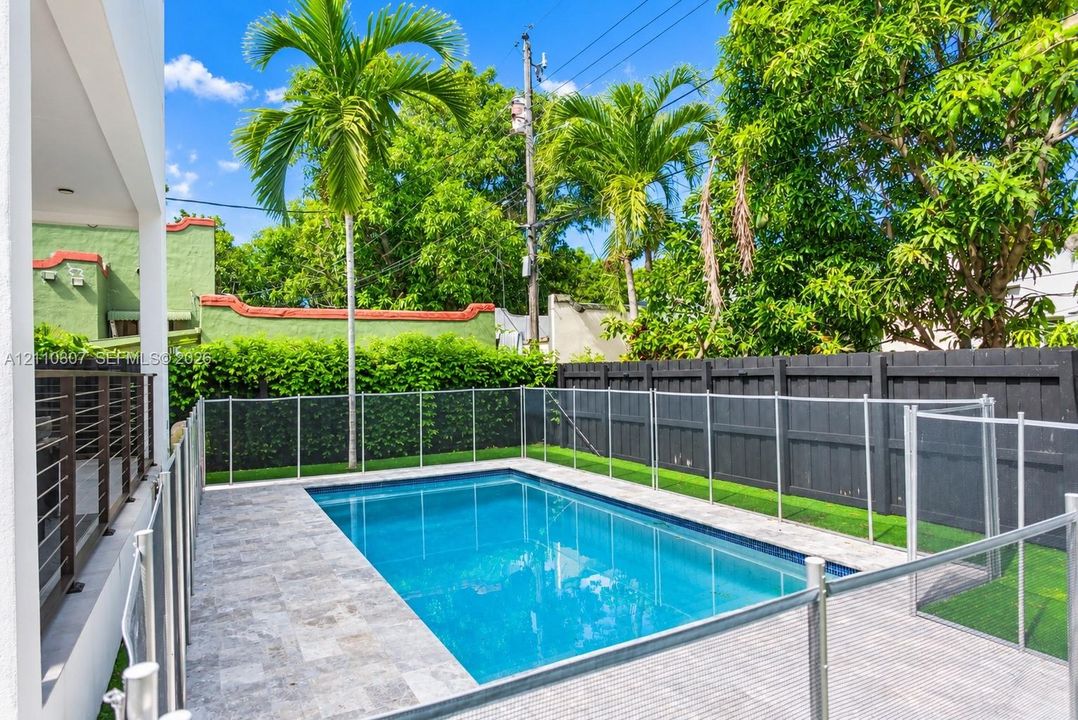 Rectangular swimming pool with natural stone decking, enclosed by a transparent safety fence, surrounded by artificial turf and dark-toned privacy fencing
