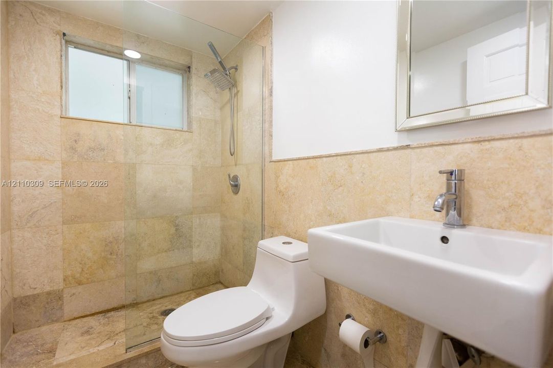 Bathroom featuring natural stone wall and floor tile, glass shower enclosure with rainfall showerhead, wall-mounted rectangular sink with chrome faucet, and mirrored medicine cabinet