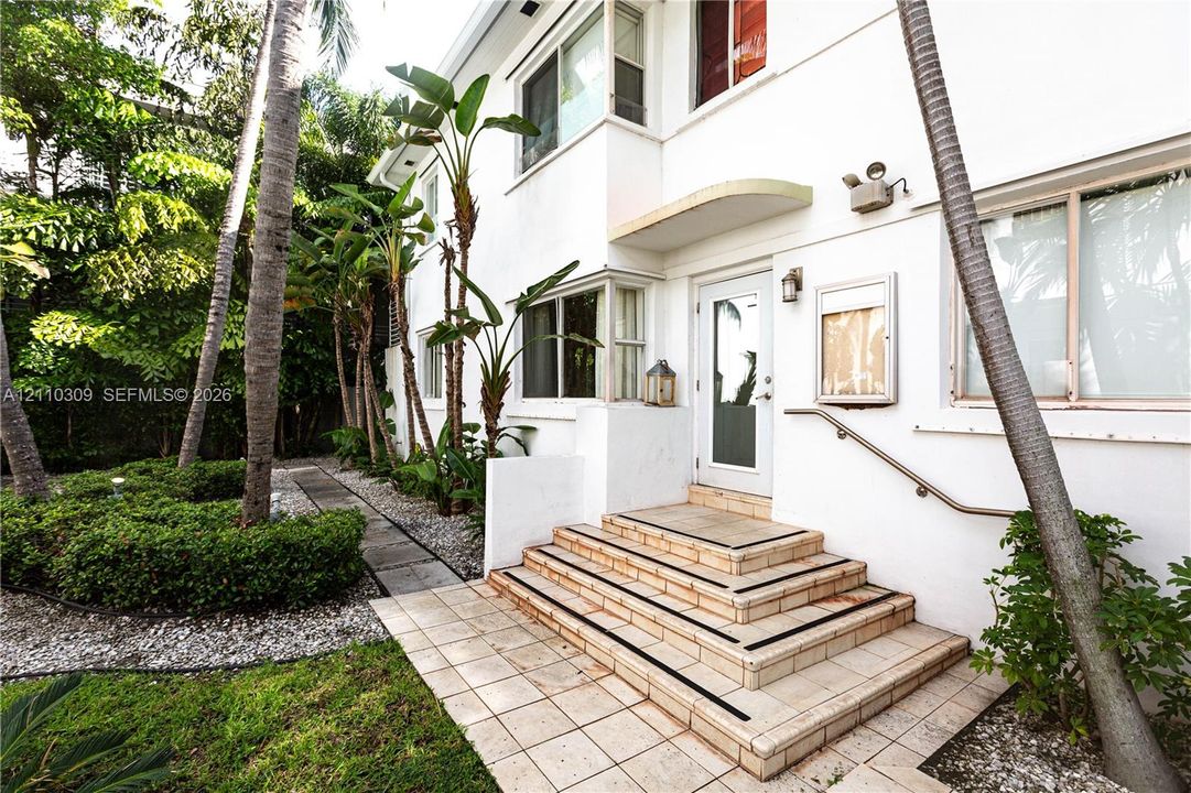 White stucco exterior featuring a tiered tile staircase, metal handrail, recessed entrance lighting, and landscaped grounds with mature palm trees