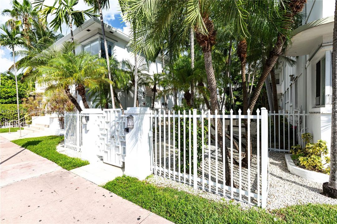 White exterior building facade featuring a gated entry, perimeter fencing, and mature palm trees