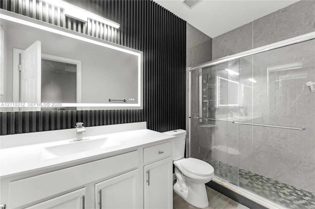 Bathroom featuring a white vanity with a rectangular sink, wall-mounted backlit mirror, black vertical slat accent wall, and a walk-in shower with grey tile surround and stone-style flooring