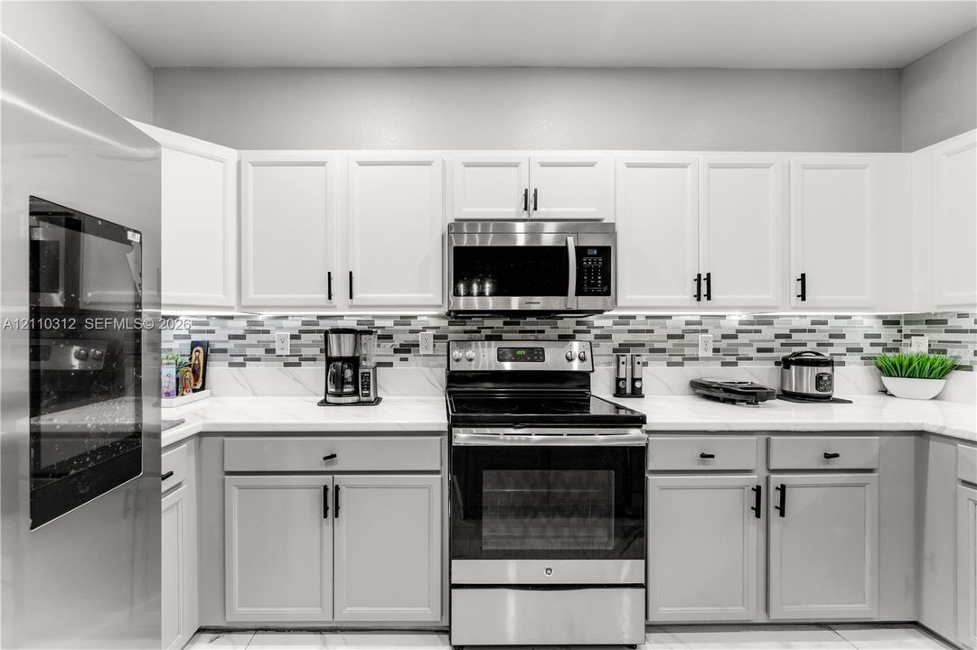 Kitchen featuring white shaker-style cabinetry, solid-surface countertops, a multi-tone glass tile backsplash, and stainless steel appliances