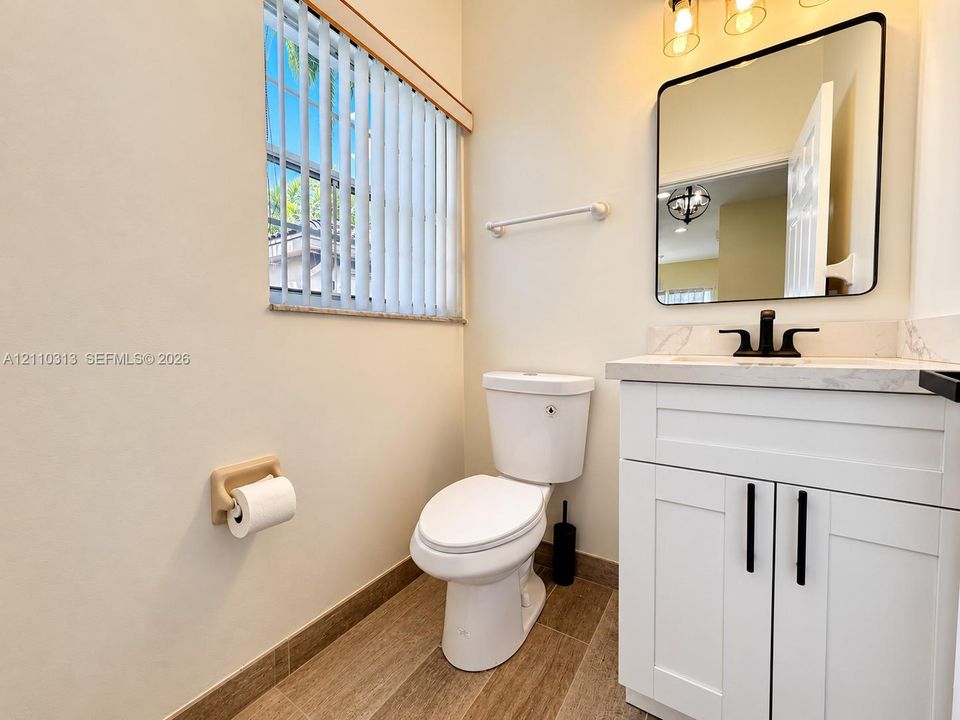 White shaker-style vanity with stone-top surface, matte black faucet, rectangular mirror with rounded corners, and wood-finish flooring
