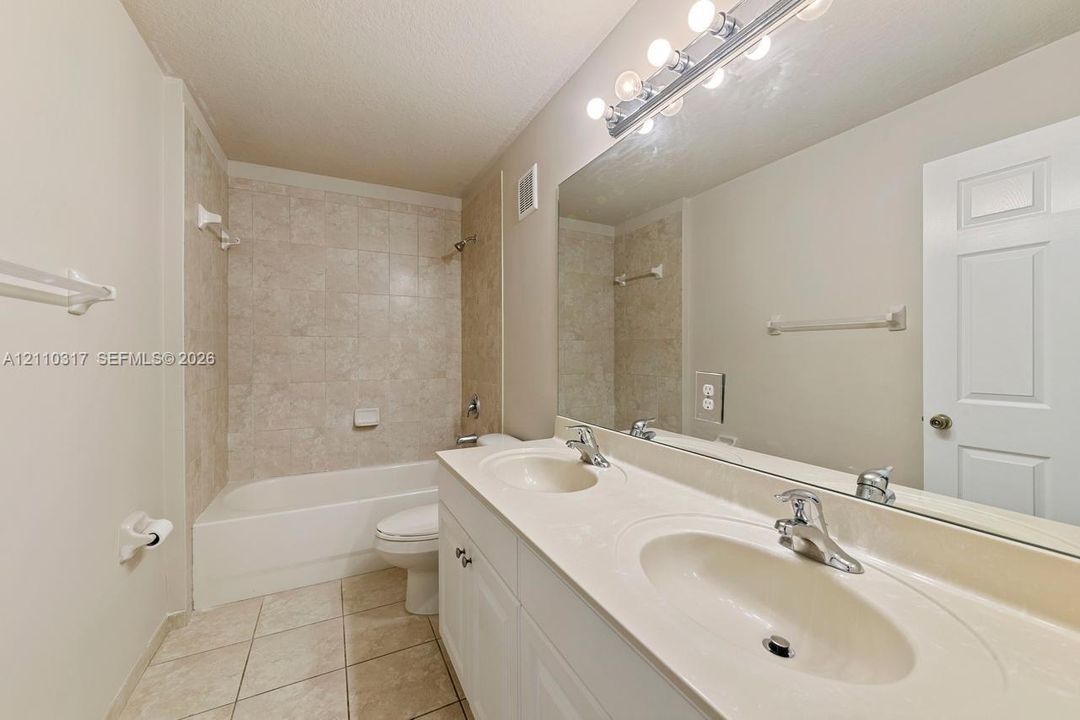 Dual-sink vanity with a solid-surface countertop, full-width wall mirror, chrome fixtures, and a tub-shower combination with tiled surround