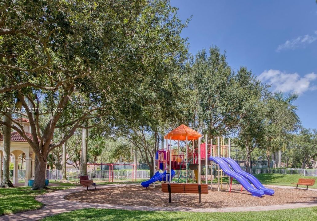 Recreational area featuring a built-in playground structure, park benches, paved walkways, and mature shade trees