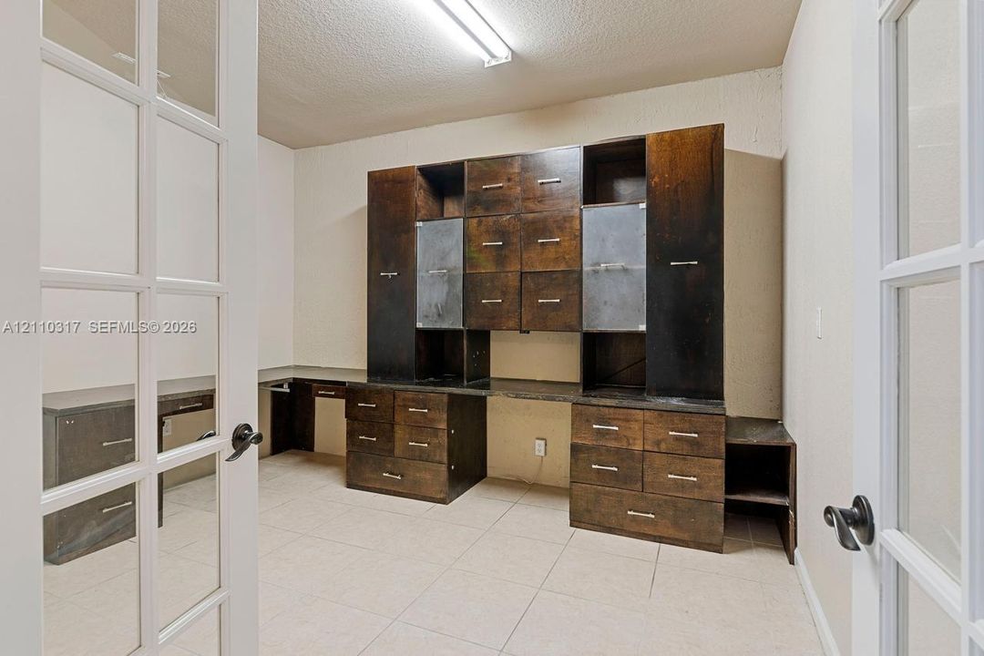 Custom built-in cabinetry with integrated desk surfaces, metal accent doors, and brushed metal hardware, enclosed by French glass panel doors and featuring ceramic tile flooring