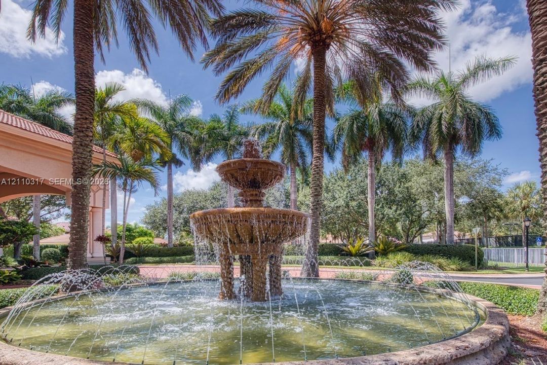 Tiered stone fountain featuring decorative architectural detailing and integrated water jets, surrounded by mature palm trees and landscaped shrubbery