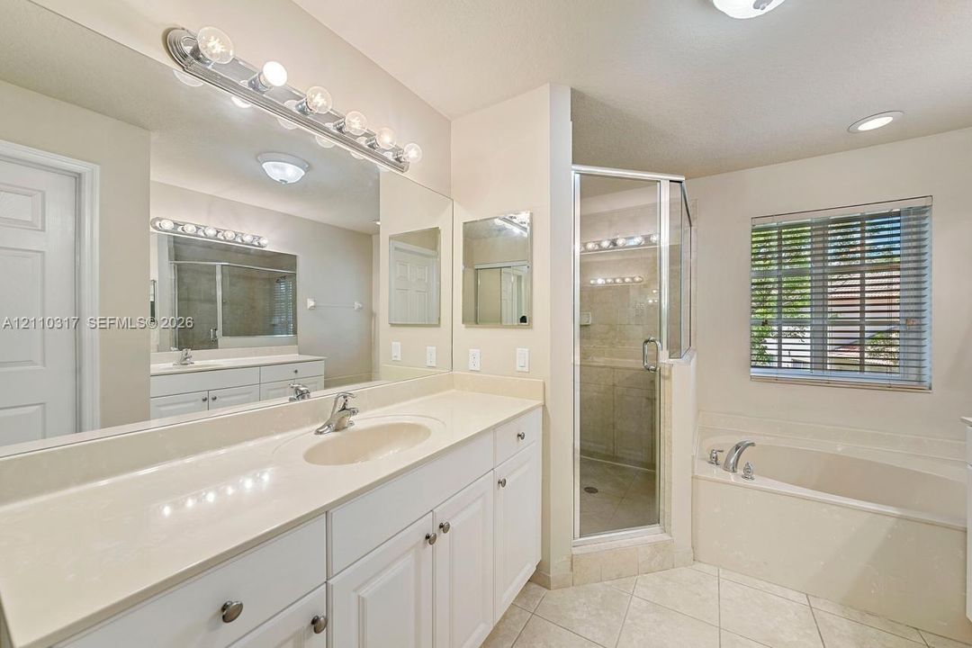 Bathroom featuring a white double-sink vanity with a solid-surface countertop, a separate glass-enclosed shower, a deep soaking tub, and ceramic tile flooring