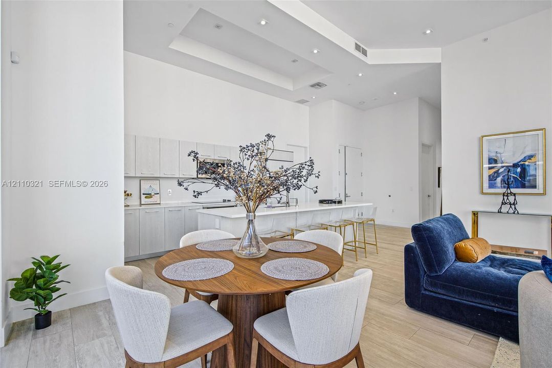 High-ceilinged open-concept space featuring recessed lighting, a tray ceiling, light-toned cabinetry, a spacious kitchen island with stone-finish countertops, and wood-finish flooring