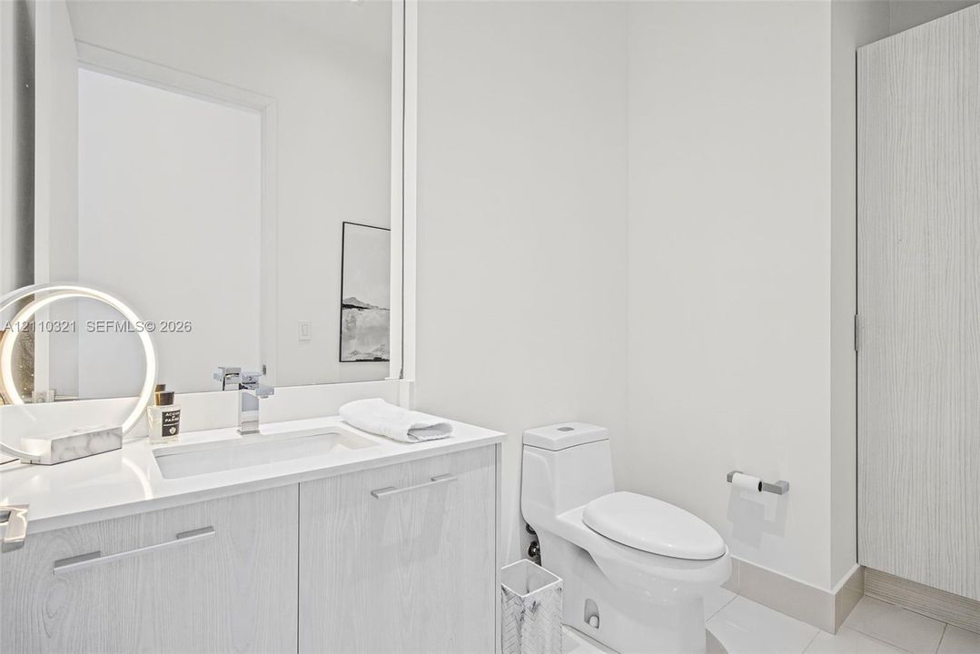 Vanity featuring a stone-surface countertop, integrated rectangular basin, chrome faucet, and wood-grain cabinetry, paired with a high-profile one-piece toilet and a full-height wall mirror