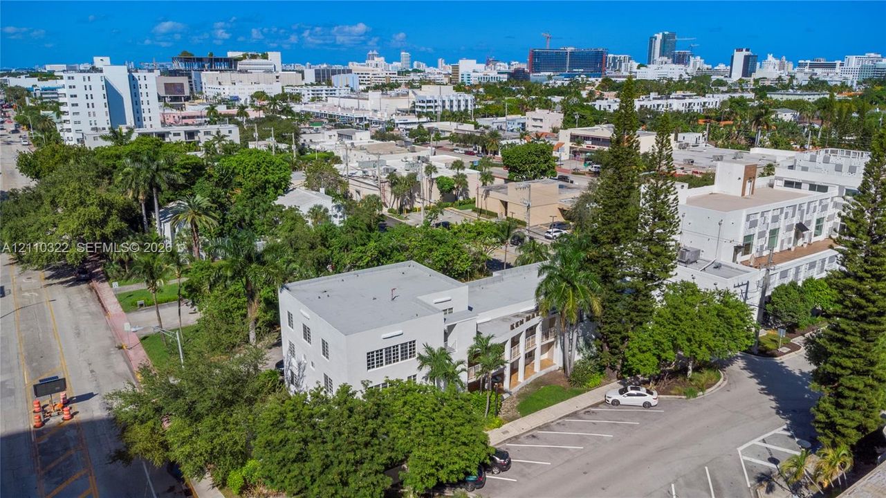 Aerial perspective of a multi-story building featuring a white facade, flat roofline, paved surface parking, and perimeter landscaping with mature trees