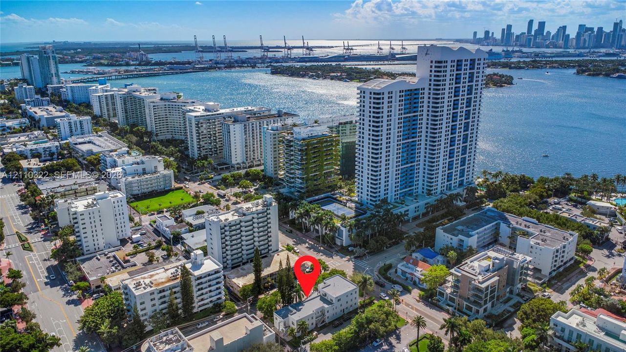 Aerial perspective featuring a low-rise multi-unit residential building with a flat roof, situated in an urban coastal setting with mature trees and proximity to waterfront access