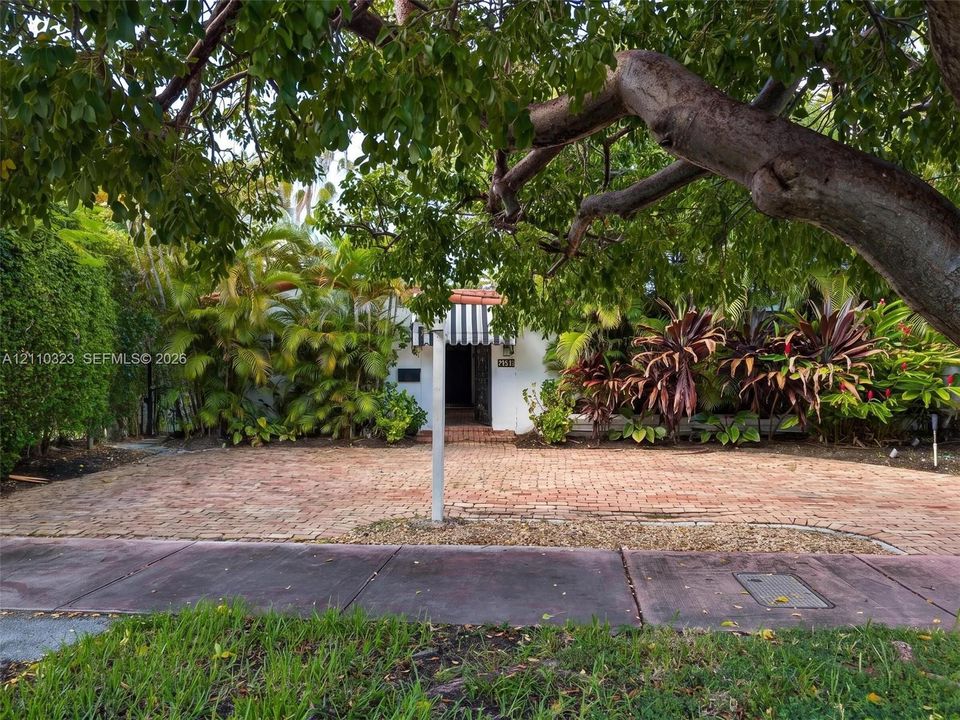 Brick-paved driveway, striped window awning, mature canopy trees, and established perimeter landscaping